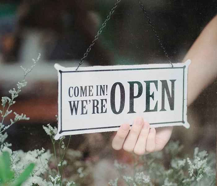 Hand flipping a sign that reads "Come In! We're Open" in a shop window